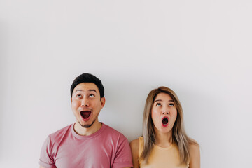 Funny shocked and surprised face of asian Thai couple lover friends, open wide mouth say wow, looking above at empty space, standing isolated on white background wall.
