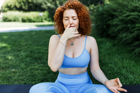 Pranayama and yoga practice outdoor, stress relief techniques. Redhead girl with curly hair sitting with crossed legs on mat in park with closed eyes, closing one nostril with thumb to reduce anxiety