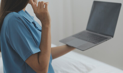 Young Asian woman with beautiful face, long hair, blue shirt lying on bed in white bedroom at home with cup of coffee and video call with laptop talking to relatives on vacation. holiday concept.