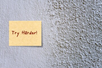 Grey painted cement concrete rough background with bright yellow note written TRY HARDER, concept...