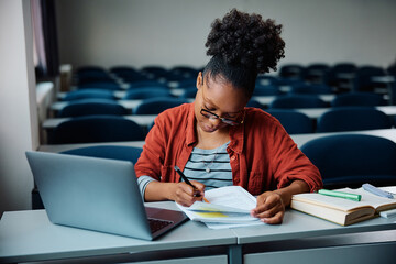 Young black woman writing in notebook while e-learning on laptop in lecture hall.