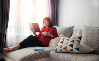  An Asian elderly woman is sitting on the sofa and reading a book, drinking some tea.