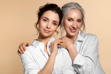 Charming beautiful mother and daughter in white casual shirt studio face forward portrait. Affectionate senior woman hugging adorable young girl shoulder from back. Adult child touching mom hand - obrazy, fototapety, plakaty