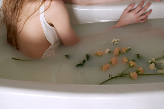 Sensual young blond woman in white swimsuit with open back sitting in bathtub filled with cloudy water and fading orange roses 
