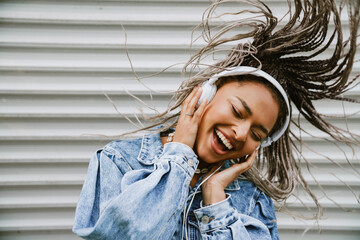 Cheerful woman dancing while listening music with headphones outdoors near gray wall