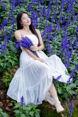 Obraz na płótnie Canvas Asian woman in white dress sitting amidst in flowers field. Beautiful girl holding bouquet flowers in hands. Happy woman enjoying in purple flower field and nature sea of fog. Embrace nature,wind.