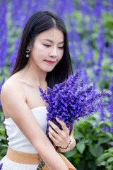 Asian woman in white dress sitting amidst in flowers field. Beautiful girl holding bouquet flowers in hands. Happy woman enjoying in purple flower field and nature sea of fog. Embrace nature,wind.