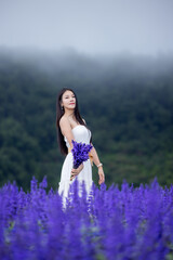 Asian woman in white dress sitting amidst in flowers field. Beautiful girl holding bouquet flowers in hands. Happy woman enjoying in purple flower field and nature sea of fog. Embrace nature,wind.