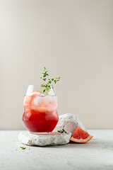 Grapefruit drink with thyme branches and ice. Glass of fruit mocktail on a stone stands. Summer cold refreshing lemonade. Food minimalism, still life. Beige background. Copy space