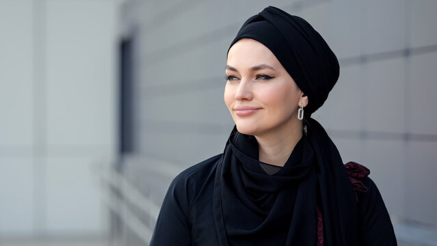 Muslim woman wearing black hijab and dark clothes walking past grey city building.