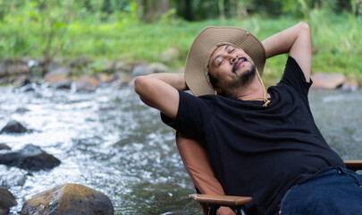 A portrait of handsome adult Asian man sleeping on a camping  chair near the stream in forest taking nap during hiking trip.