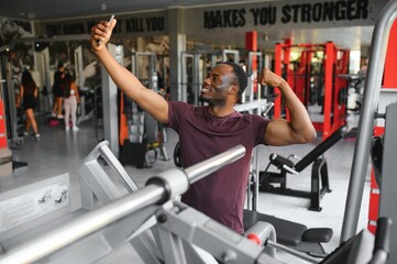 smiling young sportsman showing biceps and taking selfie with smartphone in gym.