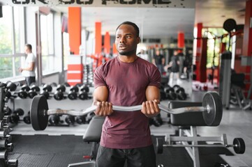 Obraz na płótnie Canvas Hot african american young man bodybuilder lifting barbell at gym, working on his arms, looking at copy space. Black muscular shirtless guy having biceps workout session. Healthy lifestyle concept.