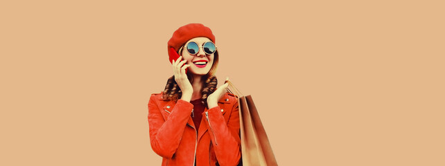 Fototapeta na wymiar Autumn portrait of happy smiling young woman with shopping bag calling on mobile phone looking away wearing red jacket, french beret hat on brown background