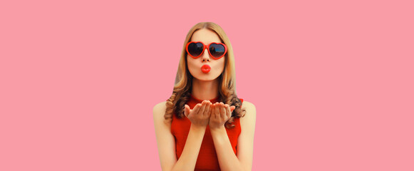 Summer portrait of beautiful young woman model blowing her lips sending sweet air kiss wearing red heart shaped sunglasses on pink background