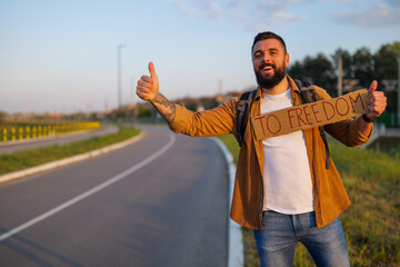 Man is hitchhiking on roadside trying to stop car. He is holding cardboard with inscription.