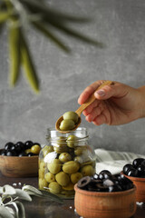 Jar and wooden bowls with olives, hand with spoon on gray background