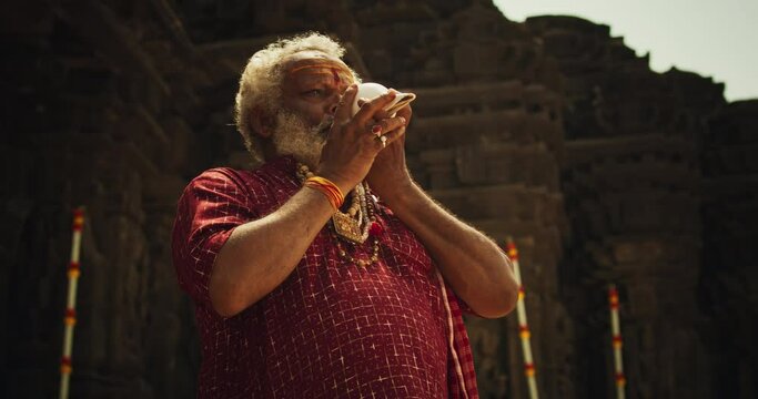 Authentic Shot of An Indian Senior Monk Blowing a Conch Shell in an Ancient Stone Temple. Elderly Man Performing Sacred Ritual on a Worship Day. Hinduism, South Asian Religion and Cultural Footage