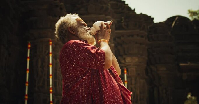 Authentic Shot of An Indian Senior Monk Blowing a Conch Shell in an Ancient Stone Temple. Elderly Man Performing Sacred Ritual on a Worship Day. Hinduism, South Asian Religion and Cultural Footage