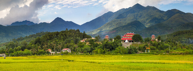 Panoramic view across rice fields of a temple on a hillside with mountains in the distance near da...