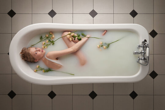 Directly above view of attractive blond woman relaxing in bathtub filled with milk and roses and looking at camera mysteriously   