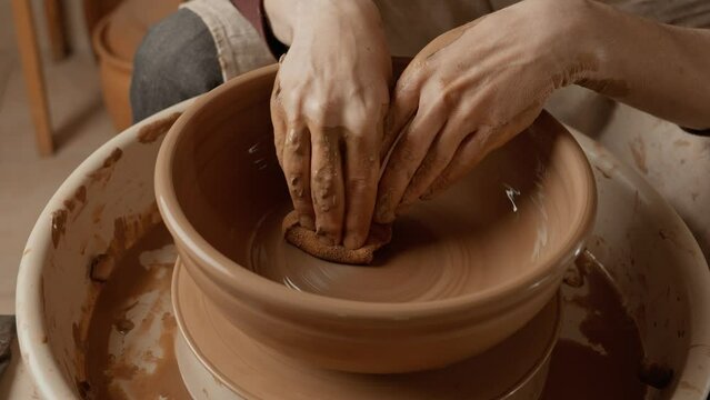 Potter making ceramic plate or dish on pottery wheel using wet sponge. Two hands create bowl. Potter shapes clay product. Close-up in 4K, UHD