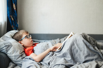 boy with glasses lies on a bed under a blanket and reads a book. A child of primary school age is...