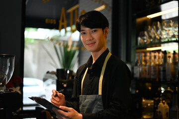 Young male waitress wearing apron standing behind counter and taking order from customer in cafe or restaurant