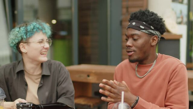 Diverse gen Z couple having fun in street cafe, young African American guy throwing french fries and catching it in his mouth while Caucasian girlfriend with prosthetic arm laughing