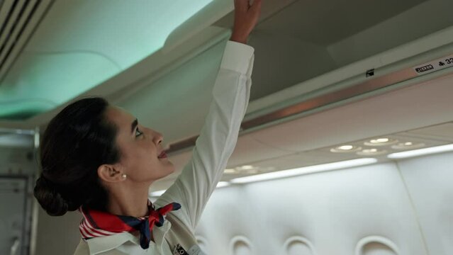 Beautiful Latina stewardess in uniform flight attendant walking to close luggage compartment and check safety seatbelt of passenger before take off working as a cabin crew position with happiness.