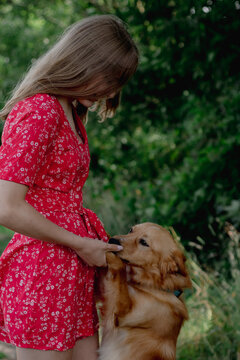 A young girl with long fair hair in a red jumpsuit hugs, strokes and plays with her dog.
