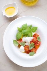 Caprese with mini mozzarella, red cherries and green basil in a white plate, vertical shot on a beige granite background, elevated view