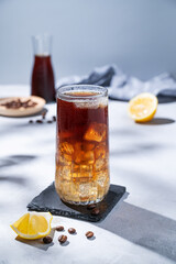 Coffee cold brew with tonic in a tall glass with ice and lemon on a light blue background with coffee beans,  bottle, fruits and shadows. Trendy summer craft refreshing drink.