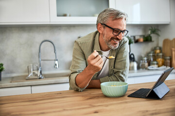 A senior man having a bowl of cereal for breakfast and using a tablet in the kitchen.