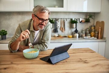 A happy senior man working over a tablet at home and having a bowl of cereal for breakfast.