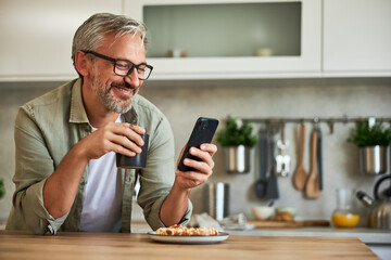 A senior man having fun on his mobile phone and drinking morning coffee at home.