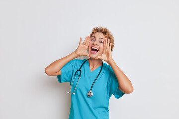 Happy female doctor posing in white studio with hands at her mouth like loud-hailer and smiling,...