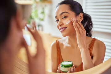 Woman applying face cream during morning routine - obrazy, fototapety, plakaty