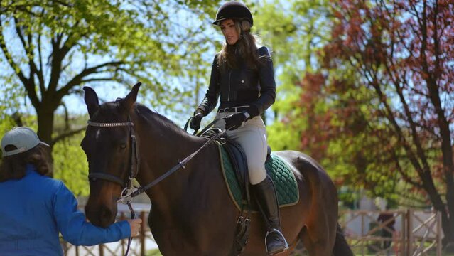 Beautiful female equestrian saddling up graceful horse with help of assistant. Portrait of gorgeous Caucasian woman mounting animal in paddock on ranch in sunshine