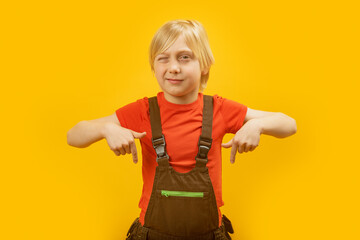 Portrait of caucasian fair-haired boy in red T-shirt and jumpsuit showing his index fingers down and winking at the camera.