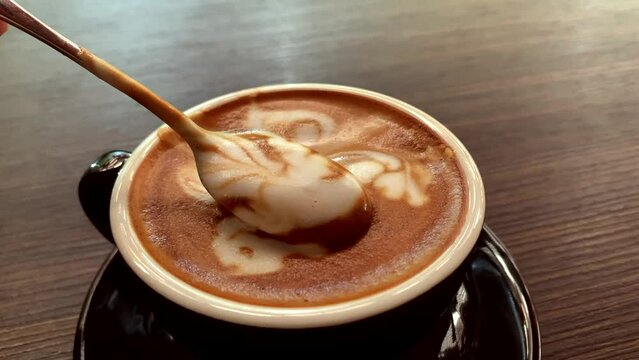 A person's hand,  no face is shown, steering the foamy cafe latte from a cup of coffee that is placed on the table.