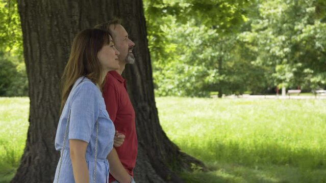 A middle-aged Caucasian couple talks and smiles as they walk through a park on a sunny day - side view