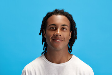 Portrait of young african man, student with dreads in white t-shirt looking at camera and smiling against blue studio background. Concept of youth, human emotions, lifestyle, fashion, ad