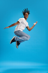 Full-length image of young african man with dreads in casual clothes jumping against blue studio background. Feeling positive. Concept of youth, human emotions, lifestyle, fashion, ad