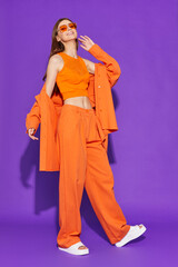 Young fashion woman in orange pants orange top orange shirt on violet background. Platform slides sandals, orange sunglasses.