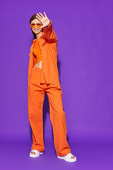 Young fashion woman in orange pants orange top orange shirt on violet background. Platform slides sandals, orange sunglasses. - 632124541