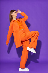 Young fashion woman in orange pants orange top orange shirt on violet background. Platform slides sandals, orange sunglasses. - 632124577