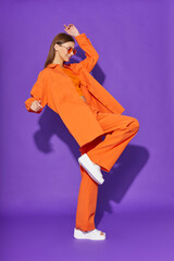 Young fashion woman in orange pants orange top orange shirt on violet background. Platform slides sandals, orange sunglasses. - 632124585