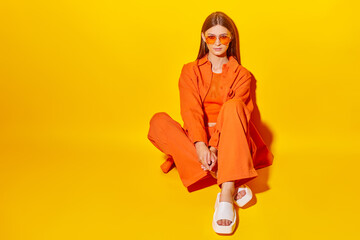 Young fashion woman in orange pants orange top orange shirt on yellow background. Platform slides sandals, orange sunglasses.