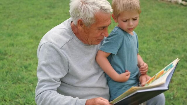 Happy grandfather reading book to curious grandson outdoors. Close up. Grandpa with grandchild spending time together. Family time comes in various forms. 4s year boy and senior man read story in park